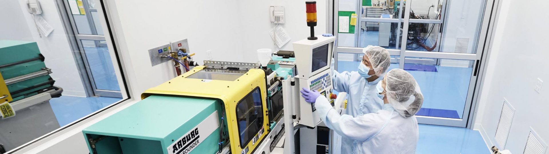 Two technicians in laboratory gear stand in front of the console of a large machine