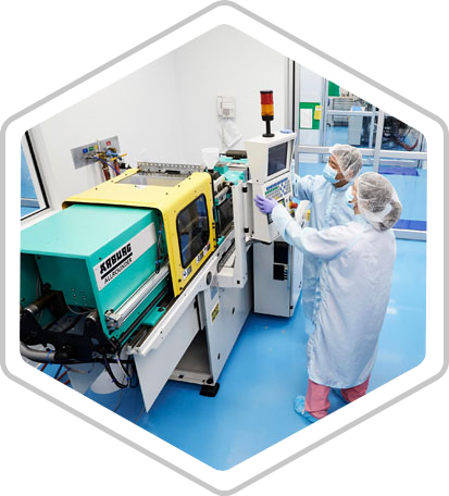 Two technicians in laboratory coats, masks and hairnets talk in front of a console on a large machine