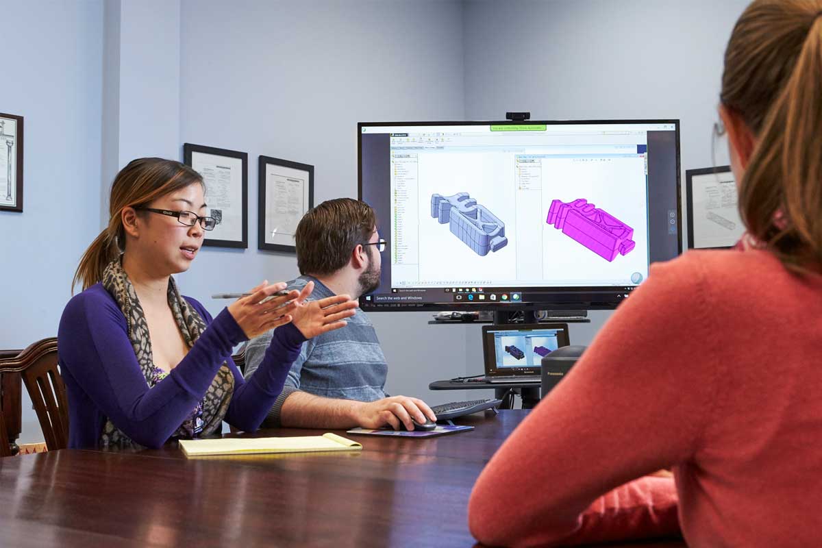 A Woman Motions With Her Hands And Talks To Colleagues In A Meeting In Front Of A Screen Showing Cad Like Drawings Of A Bioabsorbable Product